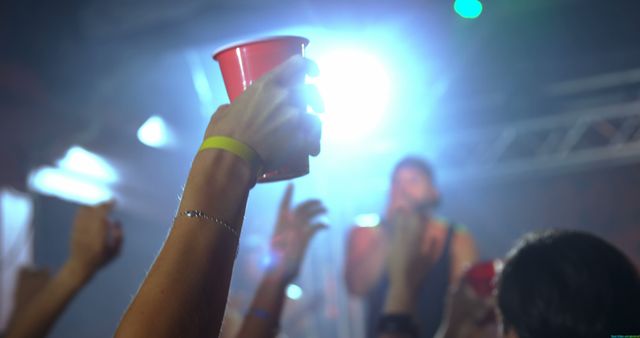A person is raising a red cup at a concert or party, with a diverse crowd and bright stage lights in the background. Capturing the energy of live events, the image conveys the excitement and social atmosphere of a music festival or club scene.