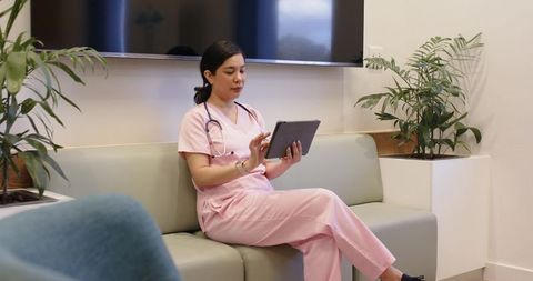 Female nurse in pink scrubs using tablet for telemedicine with stethoscope in modern clinic lobby