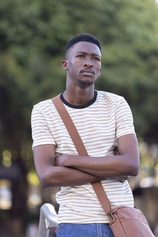 Contemplative african man standing arms crossed wearing striped tee and leather bag