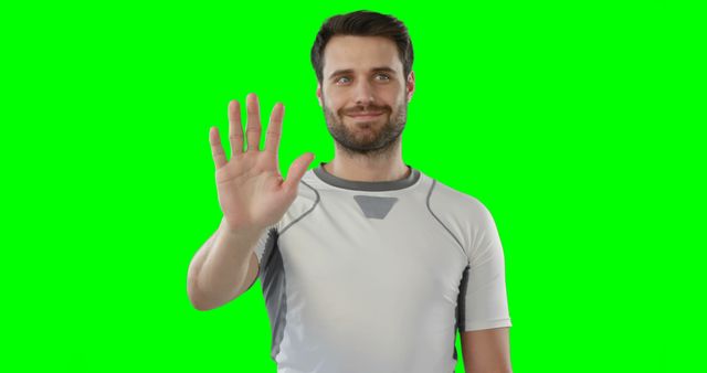 A man is waving his hand with a friendly smile on his face. He is wearing a white shirt and standing in front of a green screen background that allows for easy digital editing. This can be used for advertisements, instructional videos, or social media content where a friendly gesture is needed.