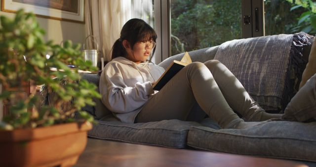 Asian girl lying on couch and reading book. at home in isolation during quarantine lockdown.