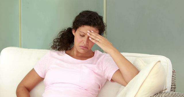 Woman in casual clothes sitting on sofa, holding her forehead in distress. Ideal for articles on health, stress management, headaches, and lifestyle. Perfect for use in blogs or advertisements related to medical care, mental health, or everyday stress relief solutions.