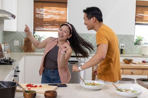 Asian couple dancing and laughing while making salad in bright modern kitchen at home