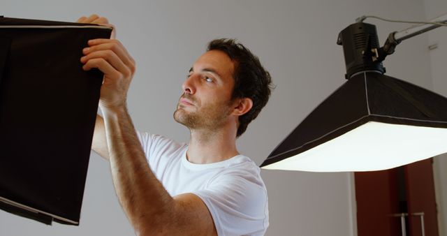 A young Caucasian man is setting up studio lighting equipment, with copy space. His focused expression suggests he is a professional photographer or videographer preparing for a shoot.