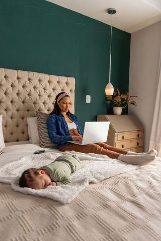 Working mother using laptop on bed with sleeping baby, home office and parenting balance