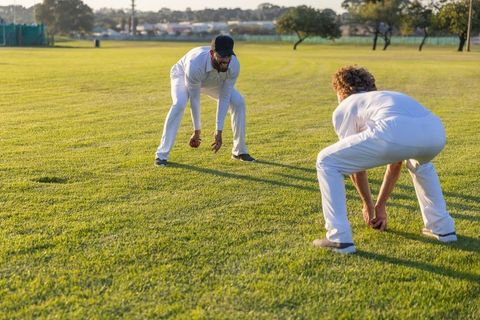 Two cricketers practicing close fielding drill on sunlit grass during training session wearing white