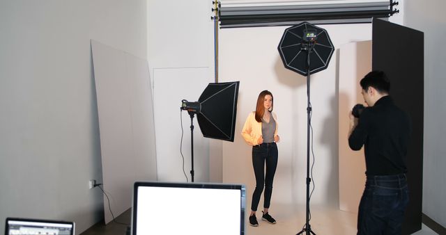 Young woman standing in a photo studio with professional lighting equipment and a photographer capturing her. This is ideal for utilizing in advertising campaigns, illustrating photography sessions, or showcasing behind-the-scenes studio work for portfolios and websites.