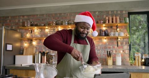 African american man whisking holiday batter in cozy rustic kitchen wearing santa hat