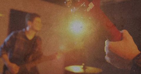 Guitarist fretting electric neck under warm spotlight with drummer blurred in background
