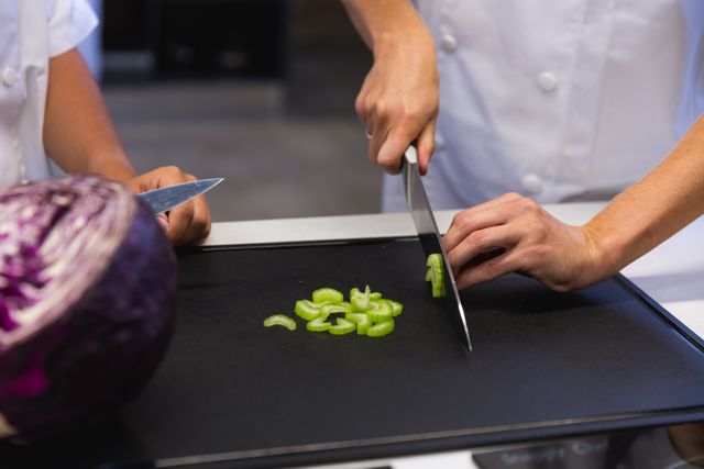Mid section of two male chefs cooking in a modern busy kitchen, slicing celery on chopping board. Cookery class at a restaurant kitchen. Workshop cooking food.
