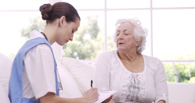 Nurse in uniform taking notes on clipboard while seated next to elderly woman in bright living room. Suitable for articles on home healthcare for seniors, medical care at home, caregiving, and elderly support services.