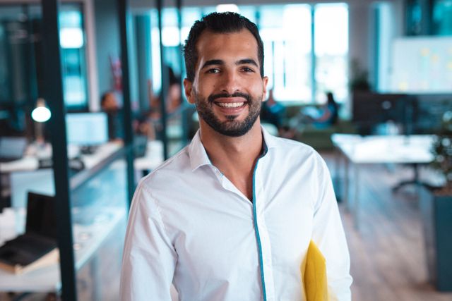 Smiling biracial male business creative standing confidently in a modern office. Ideal for use in business websites, corporate presentations, and promotional materials showcasing professional environments and young entrepreneurs.
