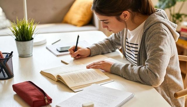Female student studying at home desk writing notes from open textbook, smartphone nearby