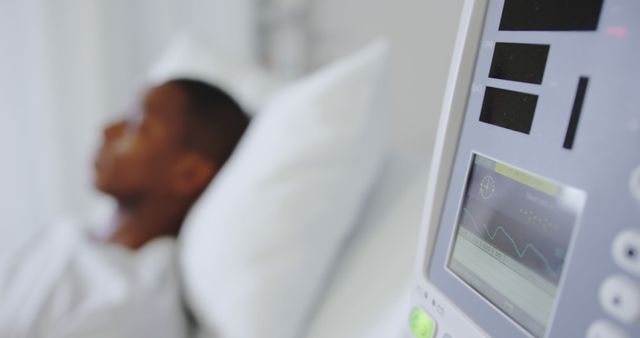 Foreground focusing on medical monitor displaying vital signs chart, with blurred background showing patient resting on hospital bed possibly convalescing. Ideal for use in healthcare marketing materials, websites discussing medical care, health insurance advertisements, and media illustrating hospitalization or medical conditions.