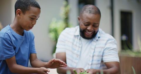 African father and son tending raised planter, teaching gardening skills on backyard patio