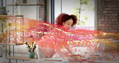 Leaning woman sorting sticky notes by laptop in loft workspace with vibrant particle stream