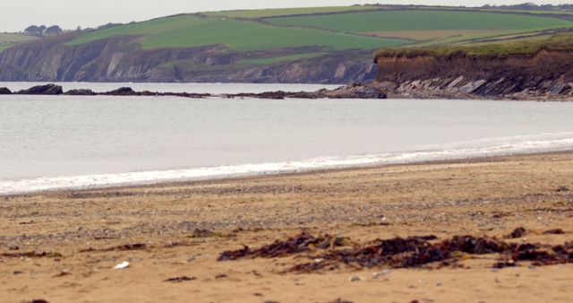 This coastal scene captures a serene sandy beach with gentle waves lapping the shore. In the background, rolling green hills and rocky cliffs add depth and natural beauty. Ideal for travel brochures, nature websites, or relaxing scenic backgrounds, showcasing the tranquility and picturesque landscapes of coastal areas.