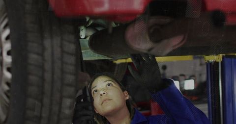 Female mechanic inspecting car underbody on hydraulic lift wearing blue coveralls and gloves
