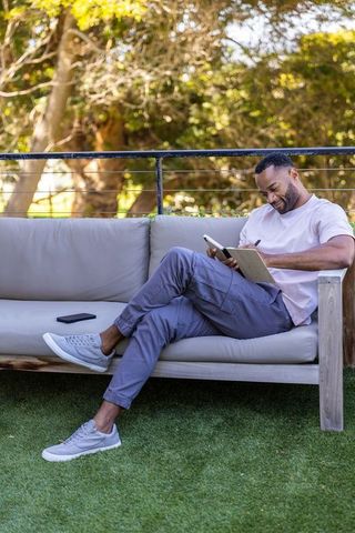Mature african american man journaling on sunlit patio sofa with phone nearby