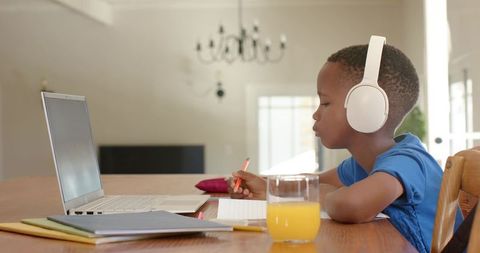 African american boy studying remotely with headphones and laptop at sunlit kitchen table