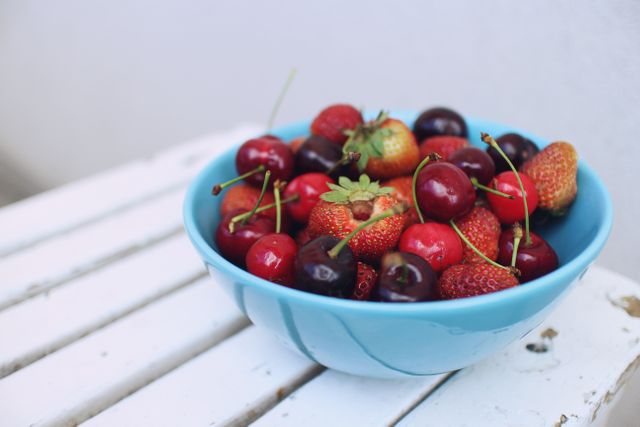 This vibrant image shows a blue bowl filled with fresh strawberries and cherries placed on a white wooden table. Perfect for illustrating concepts related to healthy eating, summer snacks, organic produce, and fresh fruit. Ideal for use in food blogs, recipe websites, nutrition articles, and promotional materials for farmers' markets or organic food companies.