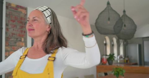 Mature woman pointing and dancing in bright kitchen wearing yellow overalls and headband