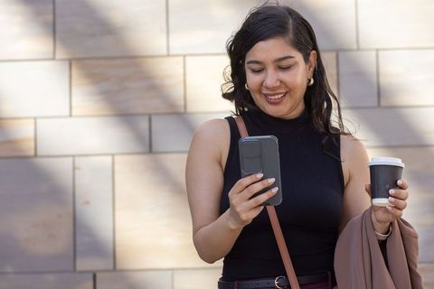 Young woman smiling while checking phone and holding coffee cup in sunlit urban plaza