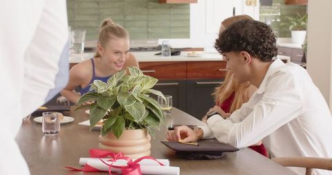 Diverse friends celebrating graduation around kitchen table with mortarboard and diplomas