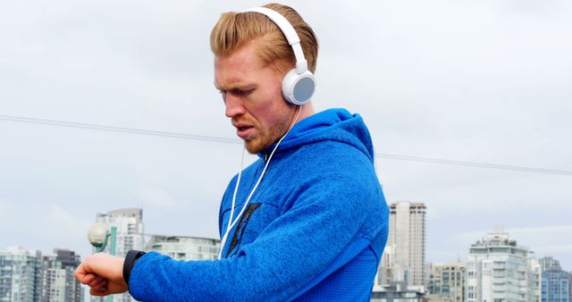 Man standing outdoors in a city environment wearing a blue hoodie and white headphones, looking at his smartwatch with a determined expression. Ideal for use in promoting fitness apps, urban lifestyle blogs, active wear advertisements, technology innovations, and health-related articles.