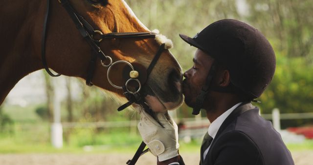 Serious african american man in riding clothes kissing horse on paddock, copy space. Sport, equestrian sports and horse riding, unaltered.