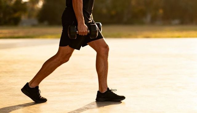 Male athlete performing dumbbell walking lunge on sunlit park path during golden hour