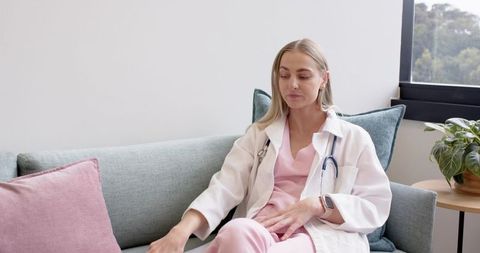 Serene female clinician resting on modern lounge sofa wearing pink scrubs and stethoscope