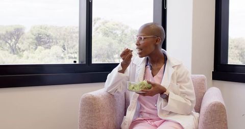 Doctor eating salad and relaxing in sunlit clinic lounge wearing pink scrubs