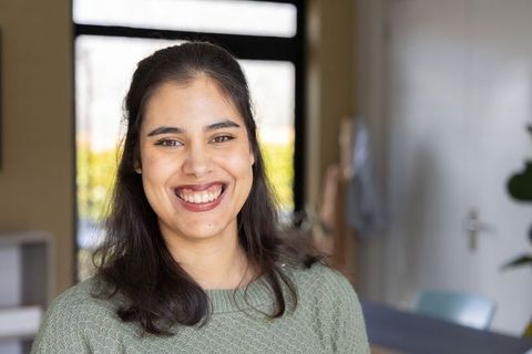 Indian woman smiling in cozy sunlit home workspace wearing green knit sweater