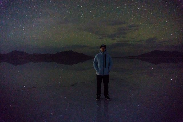 Person standing outdoors under a starry night sky with mountains in the background. They are standing on a reflective surface, likely water, which mirrors the stars, emphasizing the tranquility of nature. Useful for illustrating themes of solitude, calm, and natural beauty. Suitable for travel, nature, and lifestyle blogs, as well as meditation and mindfulness content.
