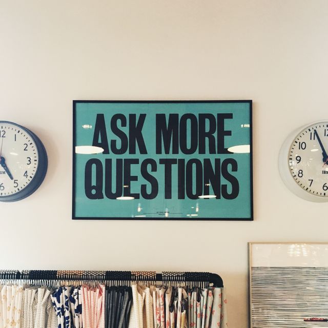 This image captures a motivational poster with 'Ask More Questions' surrounded by two wall clocks and fabric samples below. The setup offers inspiration ideal for office spaces, classrooms, and home interiors aiming for a blend of modern design and motivation.