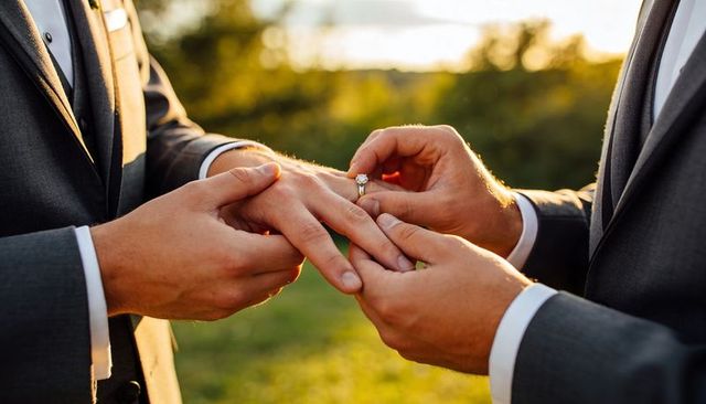 Two grooms placing diamond ring on partner's hand during golden hour outdoor ceremony