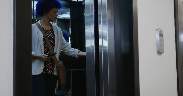 Biracial casual businesswoman with blue afro holding smartphone in office elevator. Casual office, business, communication and work, unaltered.
