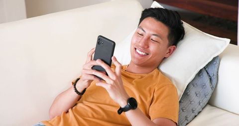 Young asian man reclining on sofa smiling while using smartphone at home for leisure