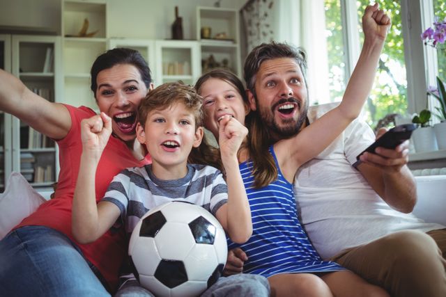 Family sitting on couch in living room, cheering and celebrating while watching a football match on television. Parents and children holding a soccer ball, showing excitement and joy. Perfect for use in advertisements, articles, or promotions related to family activities, sports events, home entertainment, and bonding moments.