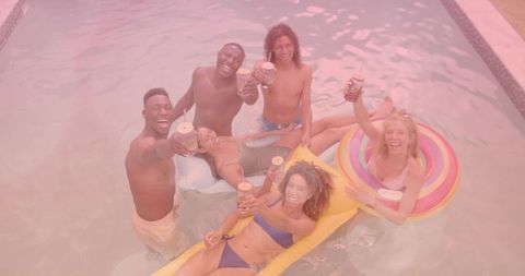 Friends toasting with canned drinks in backyard pool, laughing on colorful inflatable floats