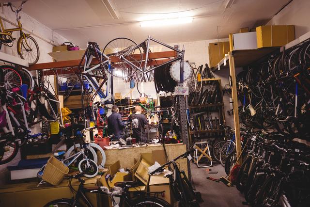 Mechanics are seen working diligently on bicycles in a busy and cluttered workshop filled with various bike parts, tools, and equipment. Perfect for depicting professional bicycle repair, teamwork in a mechanical setting, or illustrating the complexity of bike maintenance. Useful for blogs, articles on bicycle maintenance, promotional materials for bike repair services, or educational content about mechanical work.