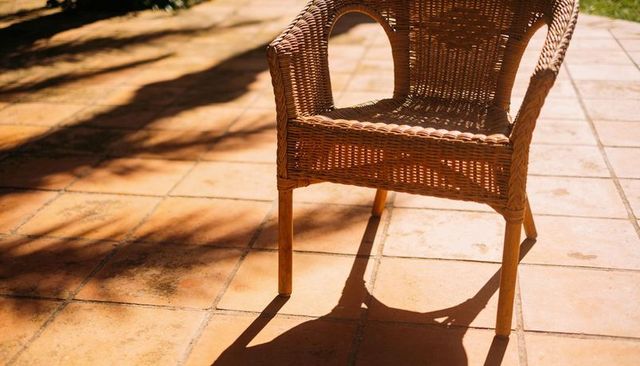 Basking wicker armchair casting long shadow on terracotta tiled patio with foliage