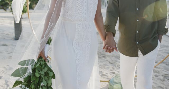 Midsection of biracial couple holding hands and flowers at wedding on beach. Love, wedding, ceremony, summer and lifestyle, unaltered.