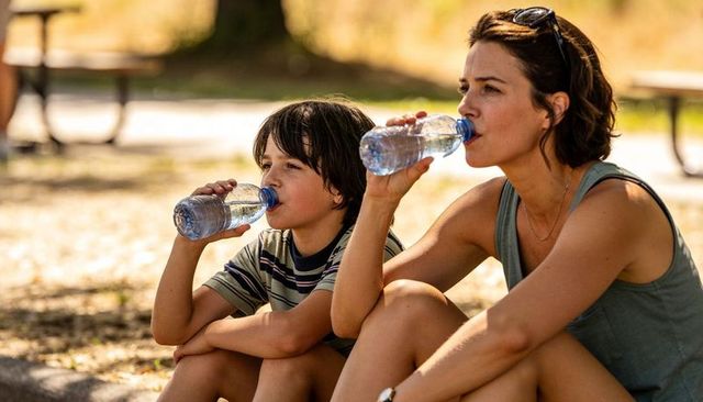 Mother and son drinking water while sitting on curb in sunlit park enjoying summer hydration