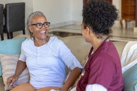 Senior african american woman smiling with caregiver on sofa providing homecare companionship