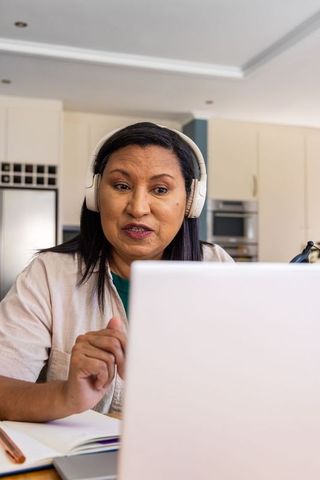 Mature woman working from home wearing headphones and using laptop with notebook