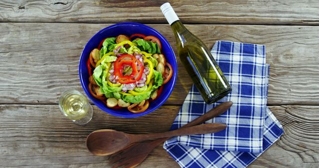 Overhead view of a fresh garden salad with a bottle and glass of white wine on a rustic wooden table. The blue bowl features a vibrant mix of freshly cut vegetables like lettuce, red pepper, and tomatoes. This scene is enhanced with wooden kitchen utensils and a blue checkered napkin, presenting a casual and inviting dining experience. Ideal for use in articles or advertisements focused on healthy eating, summer picnics, or dining aesthetics.