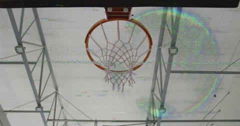 Frayed basketball hoop hanging from indoor gym ceiling with steel trusses and lens flare