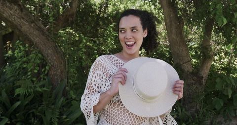 African american woman holding straw hat wearing white crochet cover-up in sunlit garden